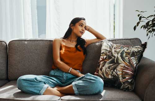 A young woman sits cross-legged on a couch. Her head is leaning on her raised arm as she looks thoughtful.