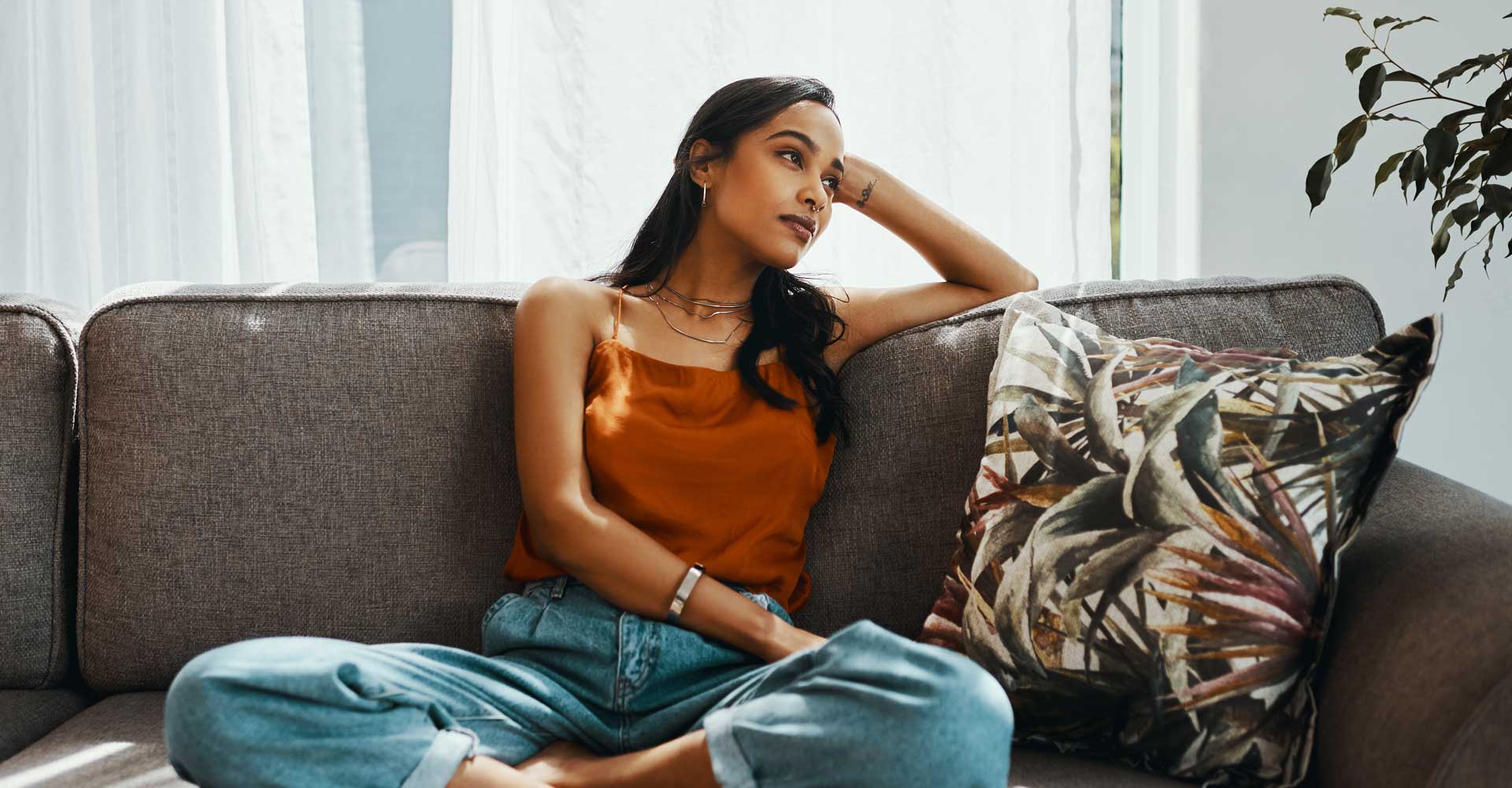 A young woman sits cross-legged on a couch. Her head is leaning on her raised arm as she looks thoughtful.