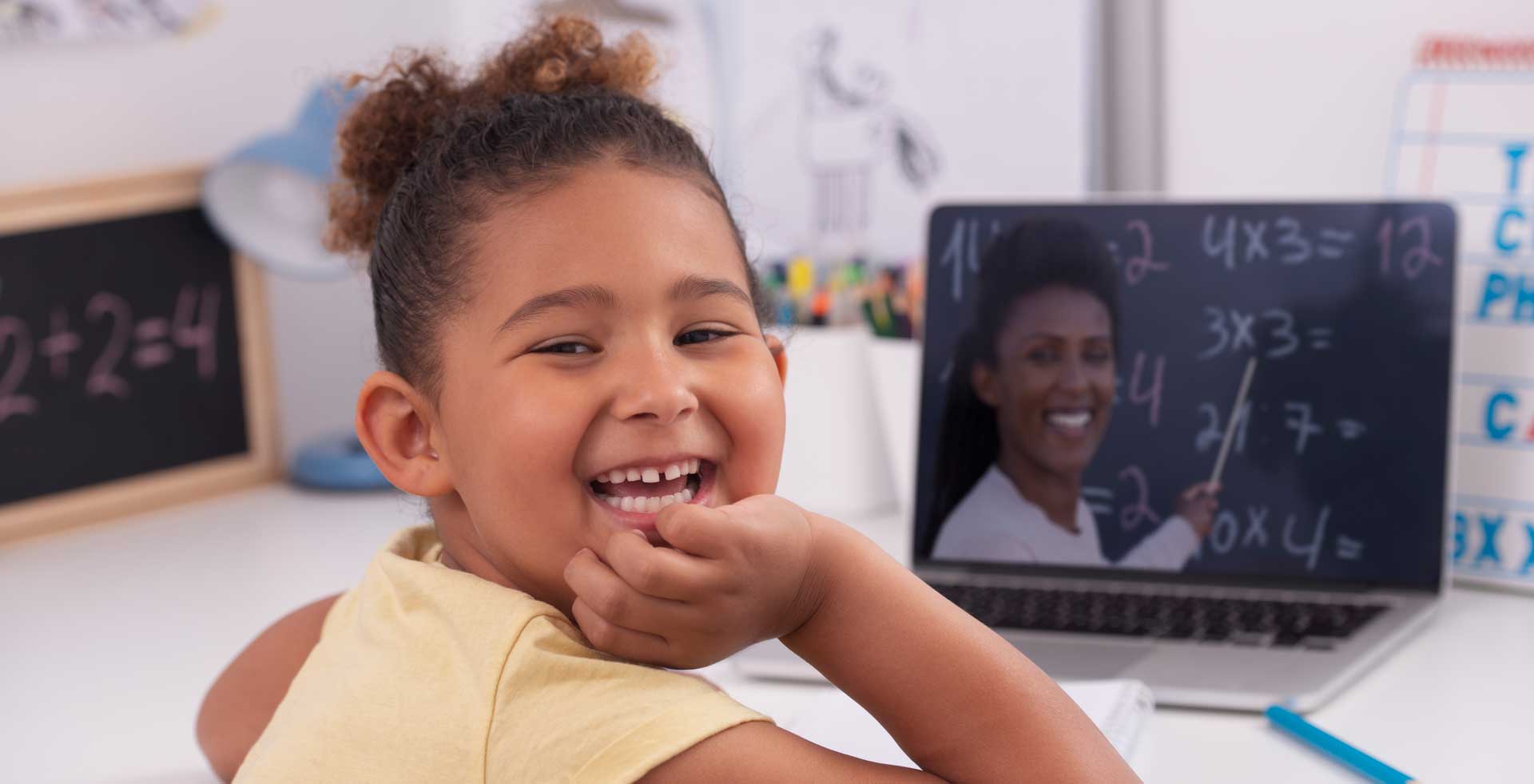 A young girl smiles at the camera, whilst she sits at a laptop that is showing a video teaching maths