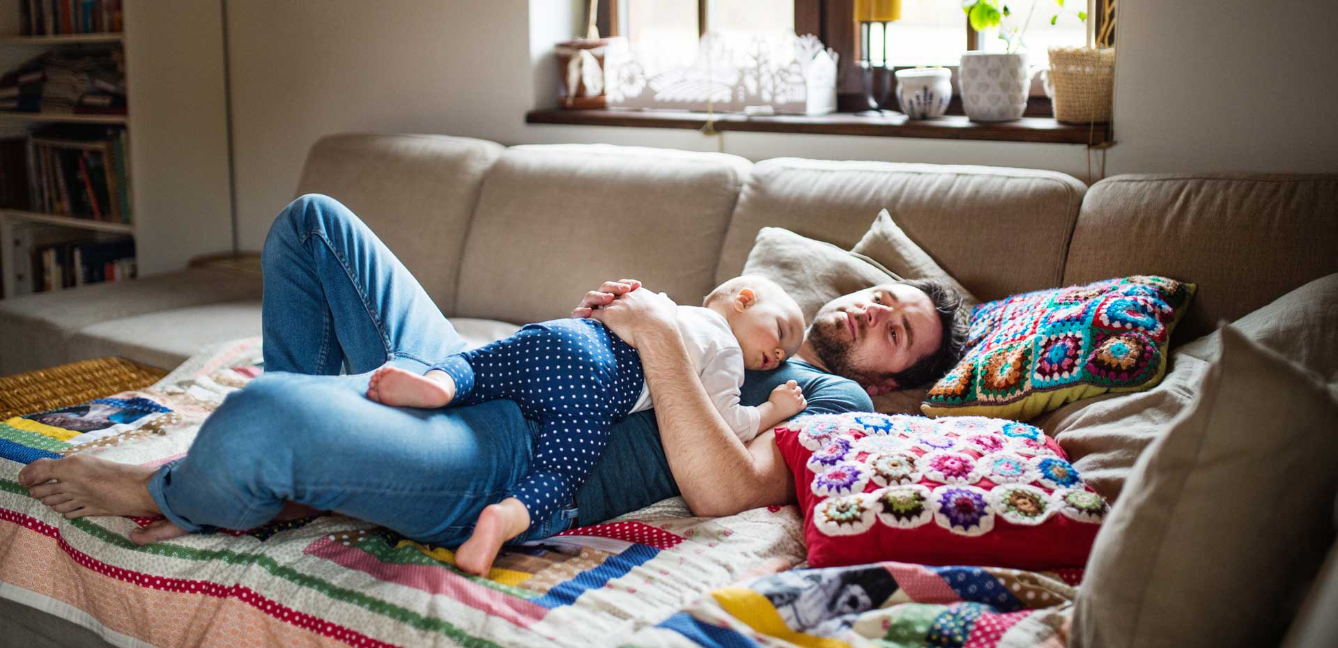 A man laying on a sofa, with his baby asleep on him.