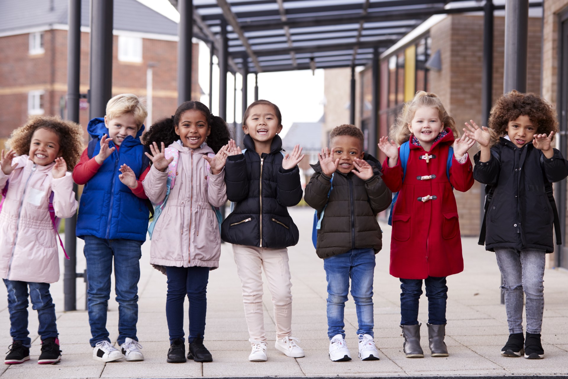 A group of smiling young multi-ethnic children standing in a row outside their nursery waving to the camera.