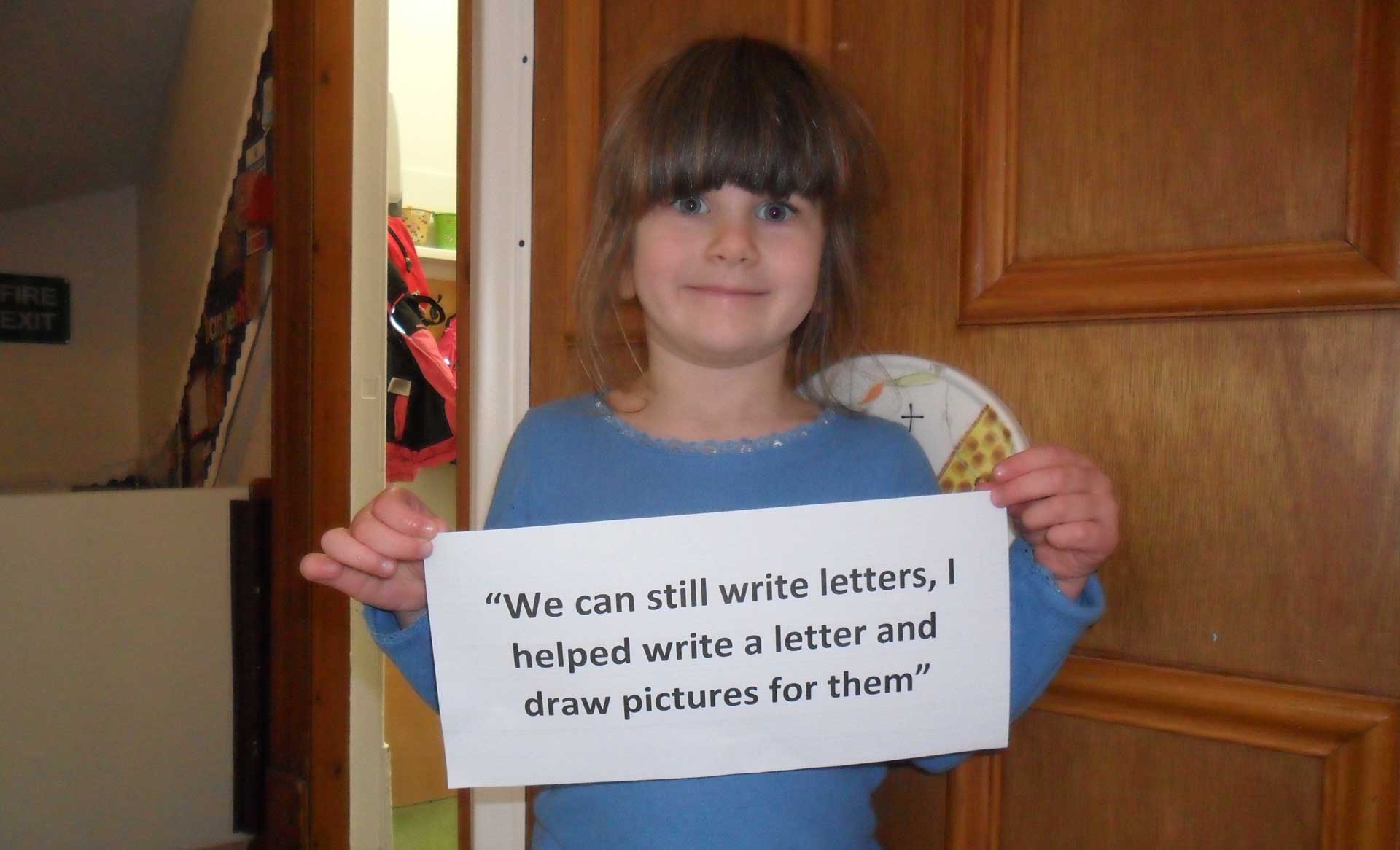 A preschool child holds up a typed note that says "We can still write letters, I helped write a letter and draw pictures for them".