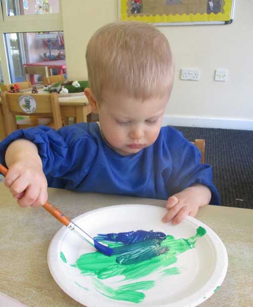 A toddler is painting a paper plate in blue and green