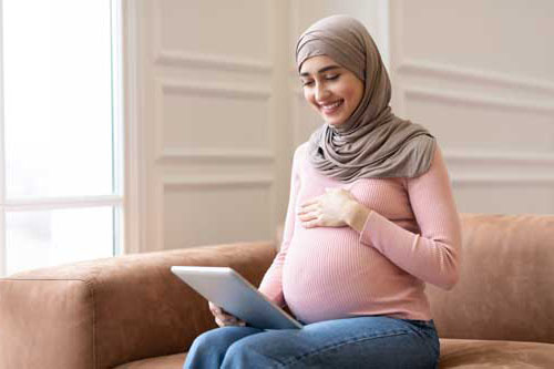 A pregnant lady sits on a sofa, looking at her tablet whilst clutching her bump
