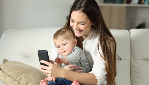 Parent holding toddler and looking at their phone with a smile whilst the child points to the phone