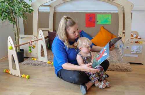 A nursery worker is sitting on the floor, reading to a child in her lap