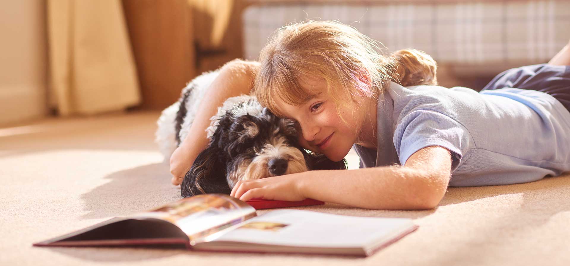 A little girl lies on the floor reading a book whilst cuddling her dog